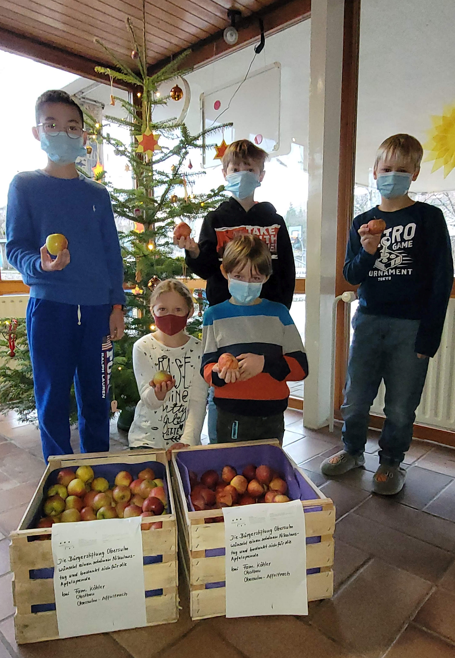 Fünf Kinder stehen mit je einem Apfel in der Hand hinter zwei gefüllten Apfelkisten vor dem Weihnachtsbaum.